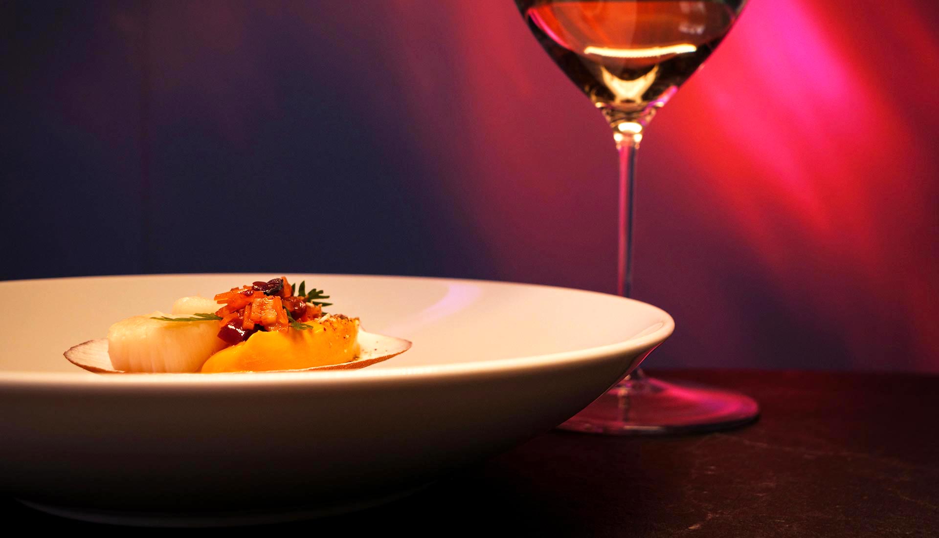 Scallop dish and glass of white wine with pink and purple visuals in the background at LUMI Restaurant